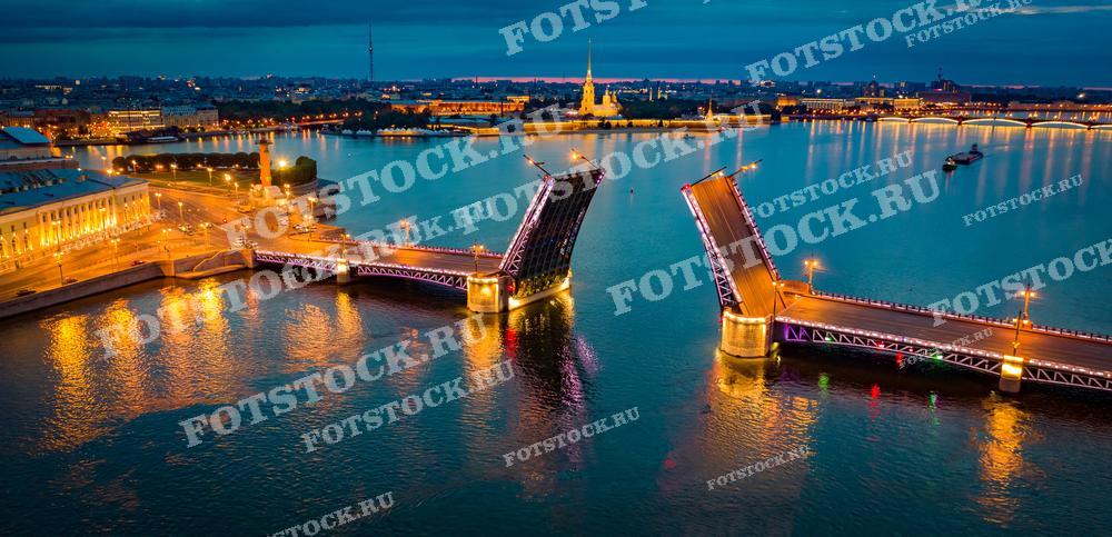 На представленной фотографии запечатлен знаменитый разводной мост через реку Неву в Санкт-Петербурге в вечернее или ночное время. Освещение моста и береговая линия выделяются на фоне сумеречного города, добавляя сцене волшебства и таинственности. Водная гладь отражает огни, что создает живописное двойное изображение моста и прибрежных огней. Этот кадр мог бы отлично подойти для туристических брошюр, постеров, а также для использования в качестве иллюстрации в книгах и статьях о культуре и истории Санкт-Петербурга.